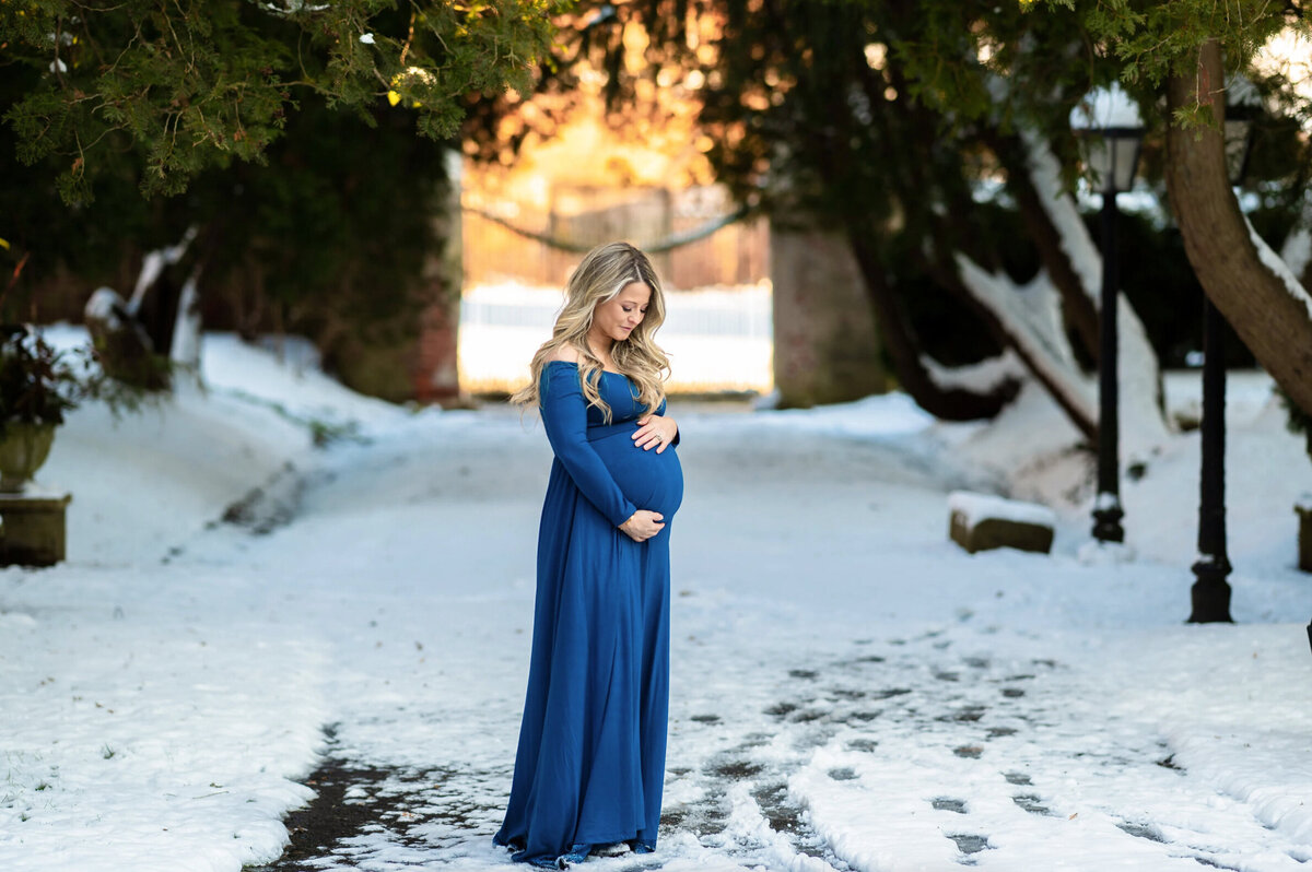Blonde woman who is pregnant, wearing a long blue dress posing for a maternity photo in the snow