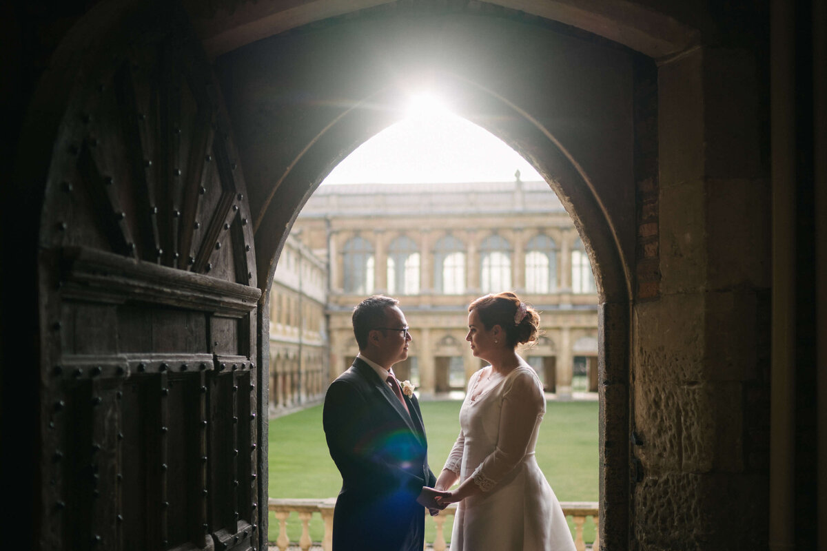 A bride and groom at a historic doorway with light pouring through from behind.