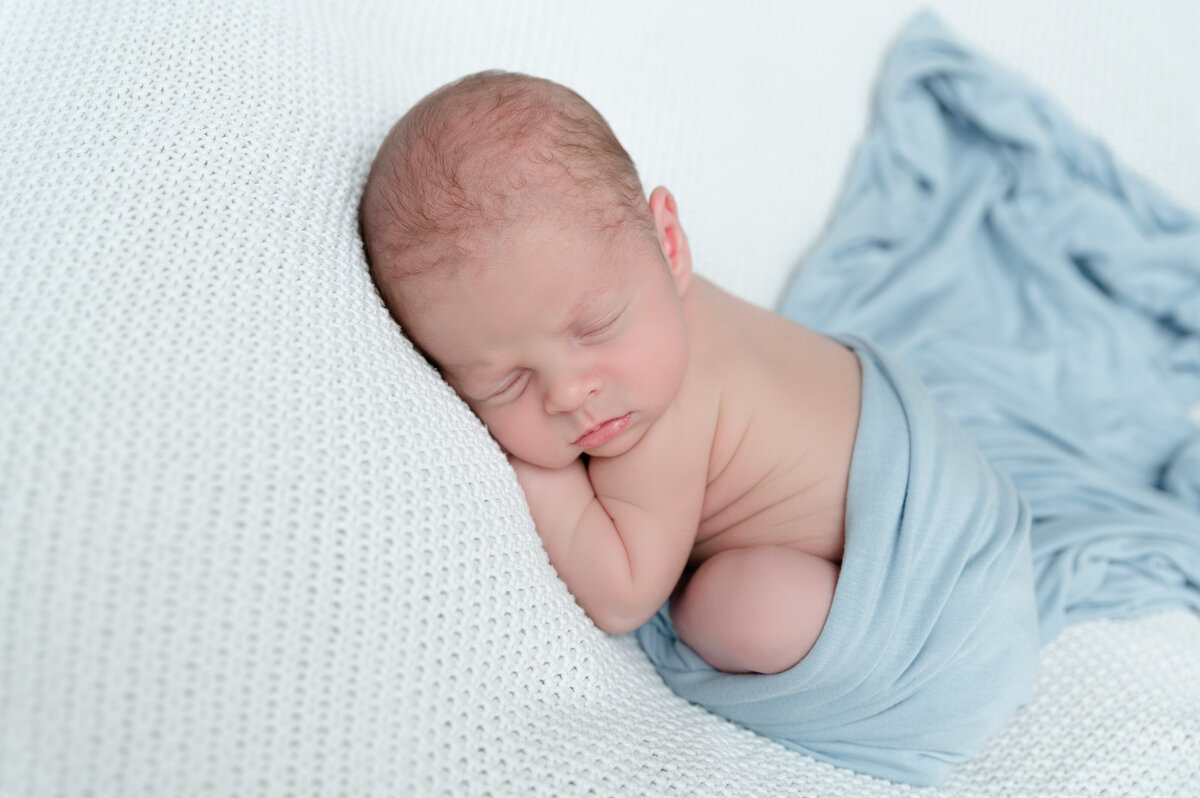 Newborn baby curled up on a blanket with a blue swaddle tucked around him.