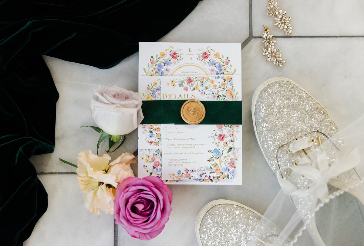 Wedding invitation with floral design, wax seal, and elegant script beside a pair of sparkly shoes, rose, and hair accessories on a marble surface at Park Farm Winery.