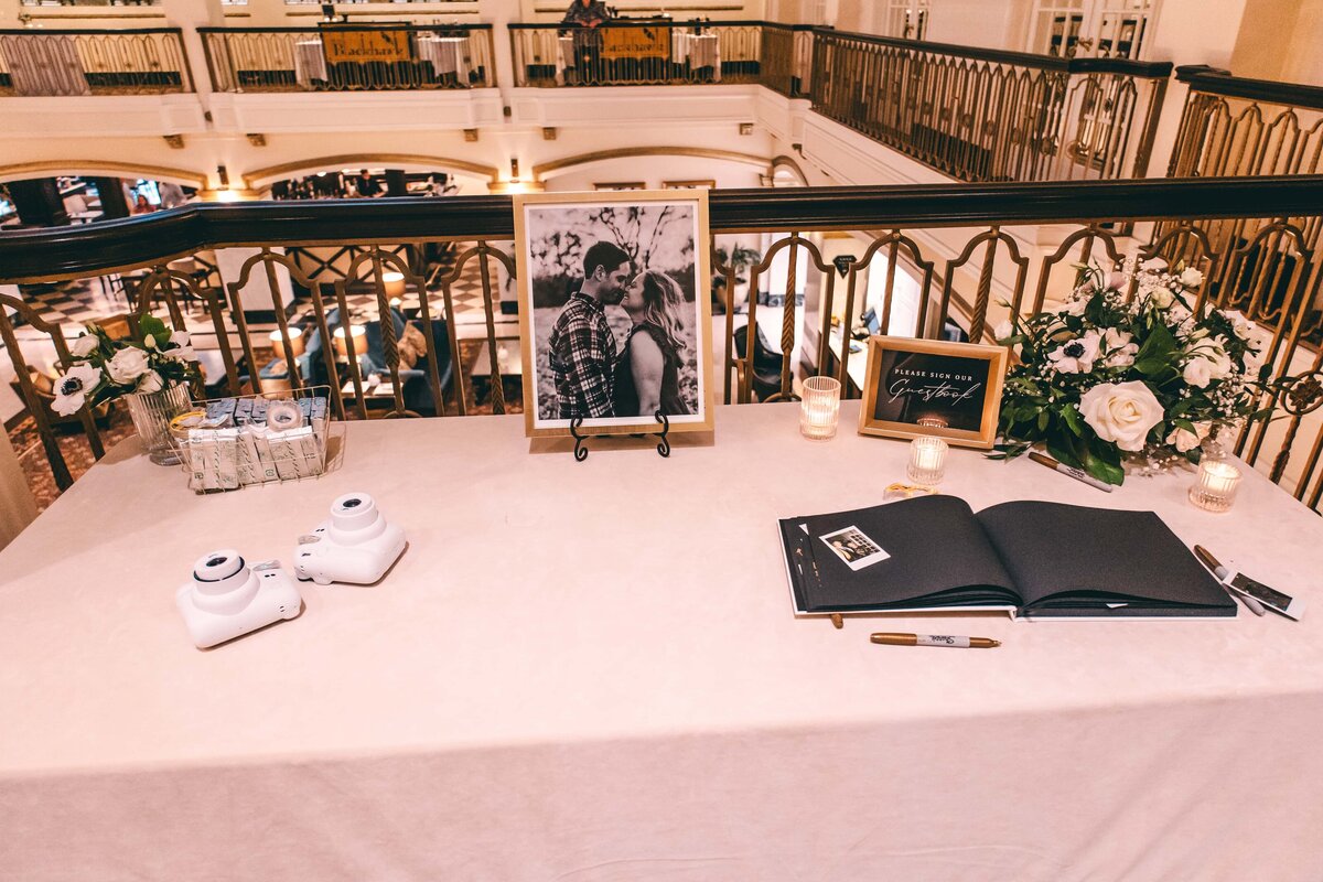 A wedding guestbook table with a framed photo of a couple, instant cameras, and floral decorations in an elegant hall in Iowa.