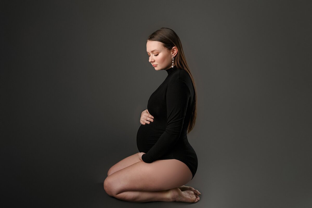 pregnant mother dressed in a black bodysuit is kneeling  on a grey background