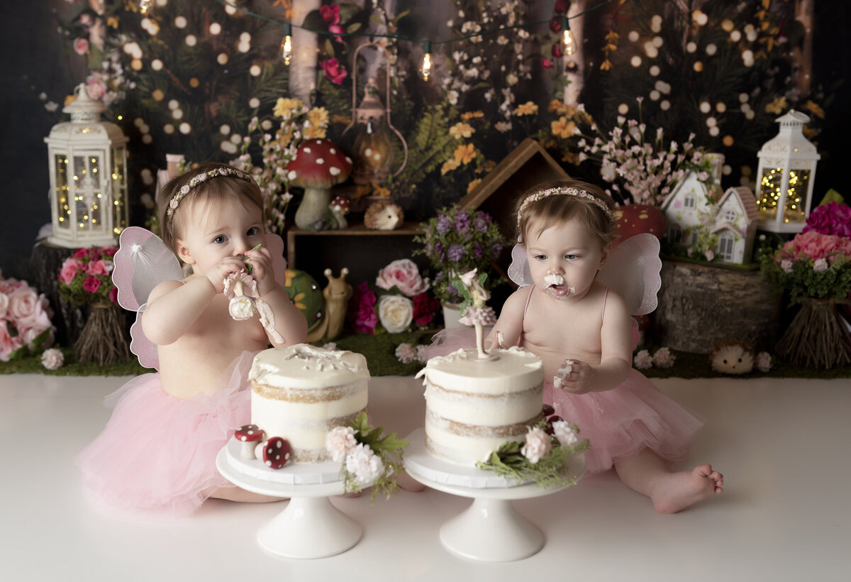 Twin girl fairy-themed cake smash in West Palm Beach, FL. Baby girls wearing pink tutus and fairy wings. The girls are eating cake and have icing on their faces. In the background there are flowers, miniature houses, mushrooms, and lanterns.