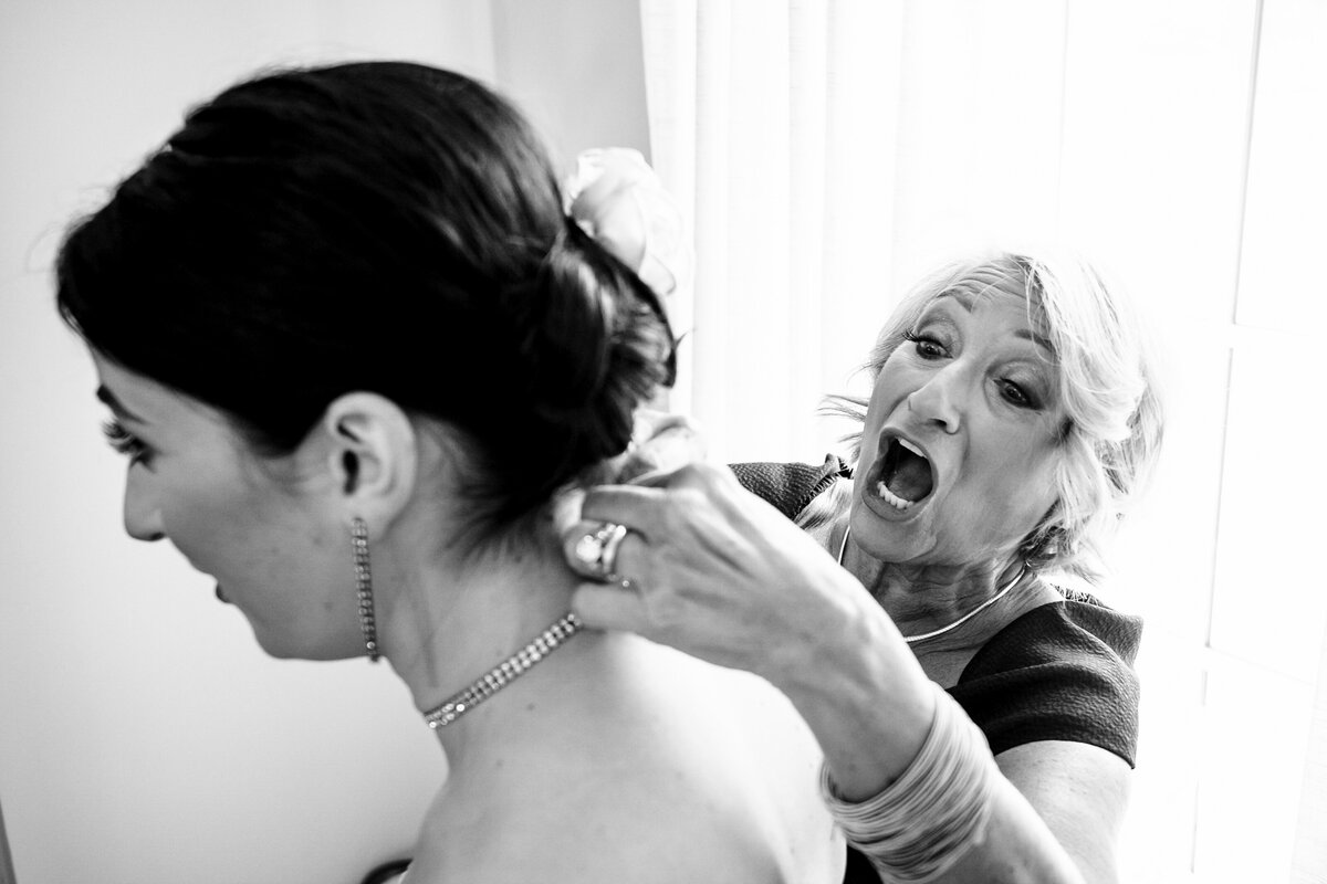 Black-and-white-candid-photograph-of-a-mother-of-the-bride-reacting-while-struggling-to-clasp-the-bride's-necklace-in-the-bridal-suite-of-Childress-Vineyards