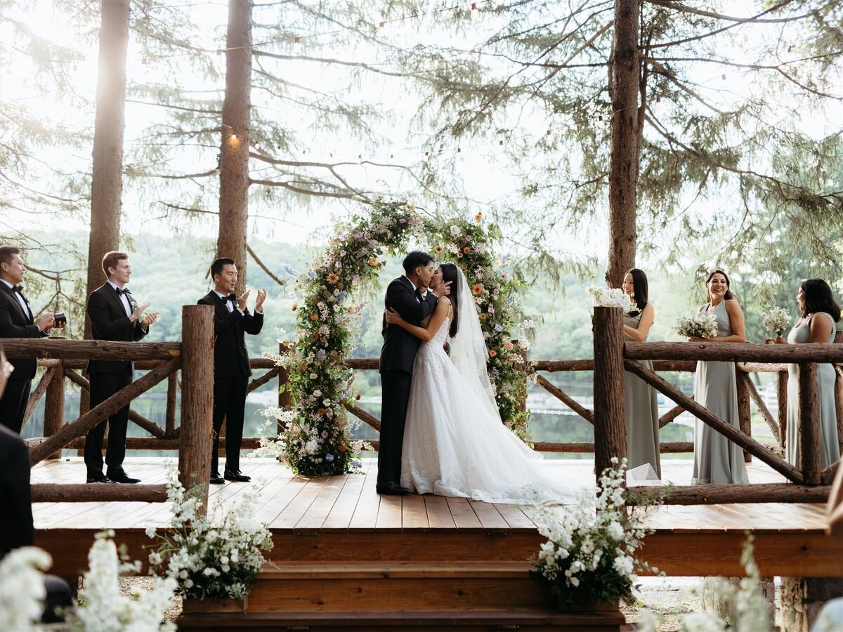 Bride and groom embrace for kiss during wedding ceremony on deck in front of stunning floral arbor, pine trees, and lake in Hudson Valley.