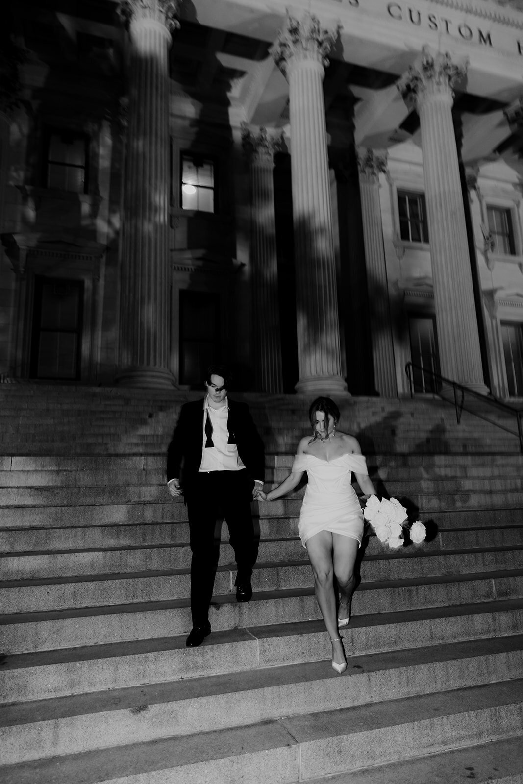 black and white photo of couple walking down stairs. Man in casual after party look with open bowtie and woman with white dress and flowers in her hand. CHarleston engagement photos by Franzi Annika
