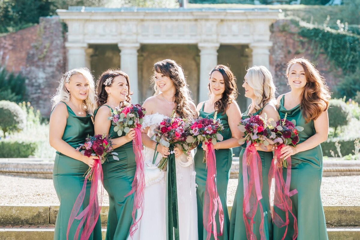 Bride and her bridesmaids, edited in a light and airy style, at Wotton House in the Surrey Hills . The bride is in white with a lovely colourful, green and pink bouqet and the bridesmaid are wearing dark green satin dresses holding bouquets and all leaning in looking at each other and smiling