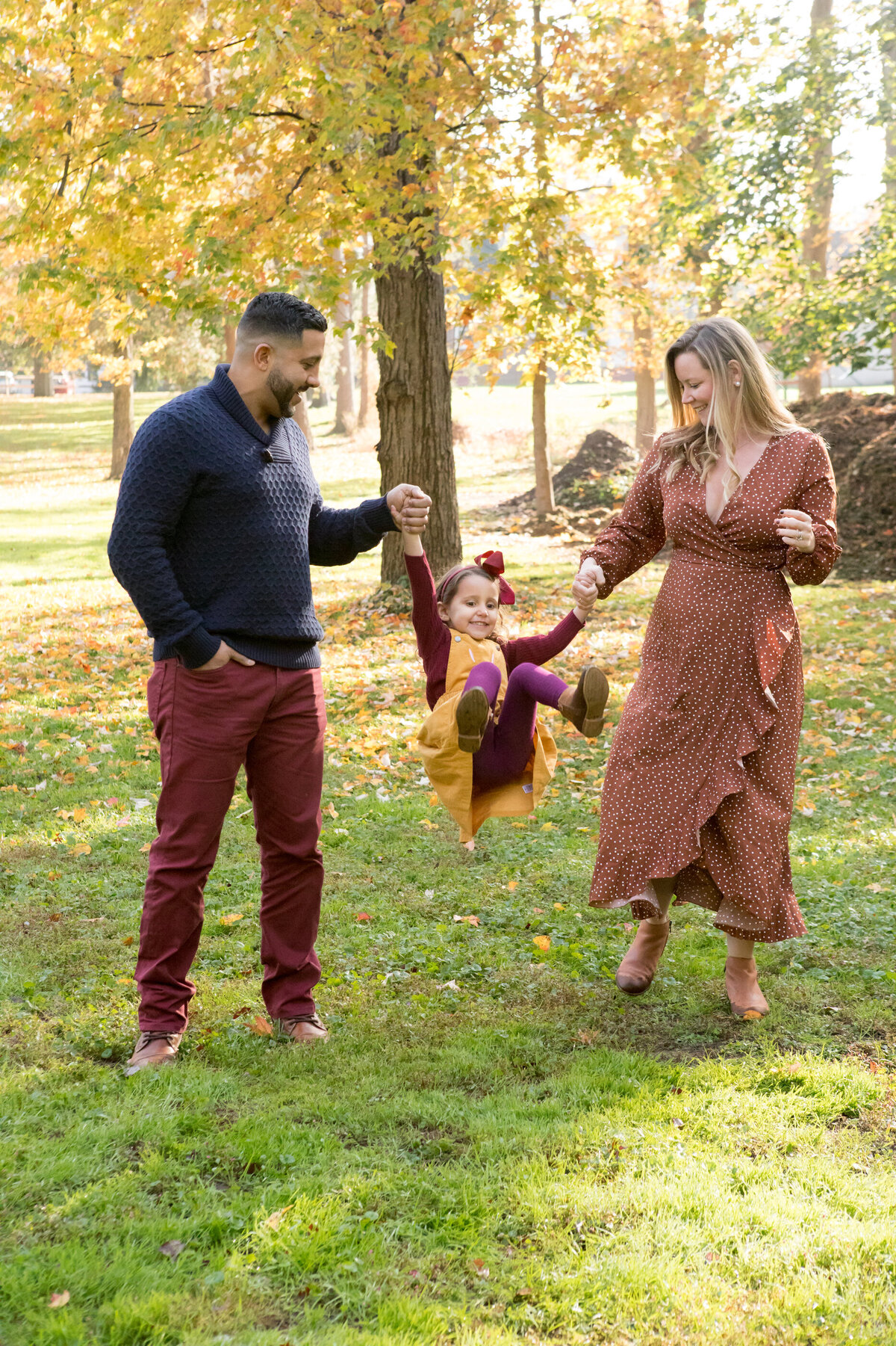 A white mother in an orange dress and a Hispanic dad in a blue sweater and maroon pants swing their young daughter in a yellow dress Hudson Valley family mini-session photographer Milestone Images