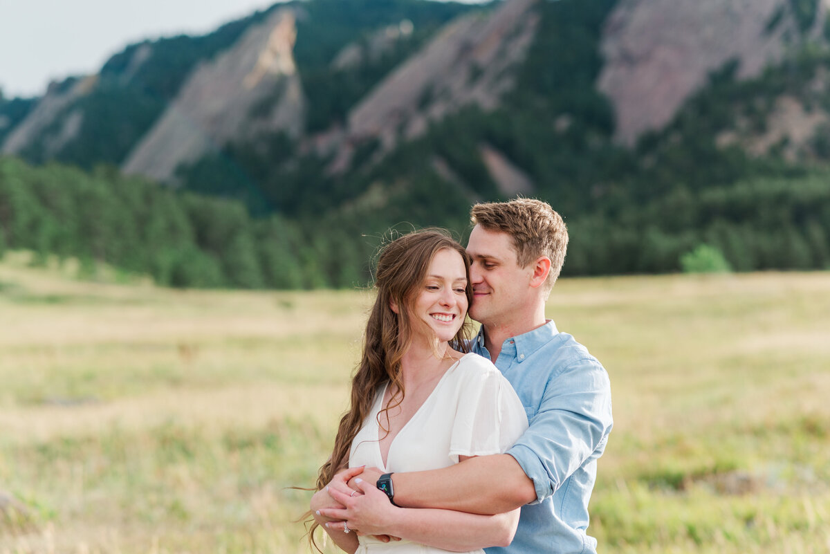 A man in a blue top hugs his fiance from behind and she looks over her shoulder toward him while he nuzzles into her ear, captured by Denver wedding photographer