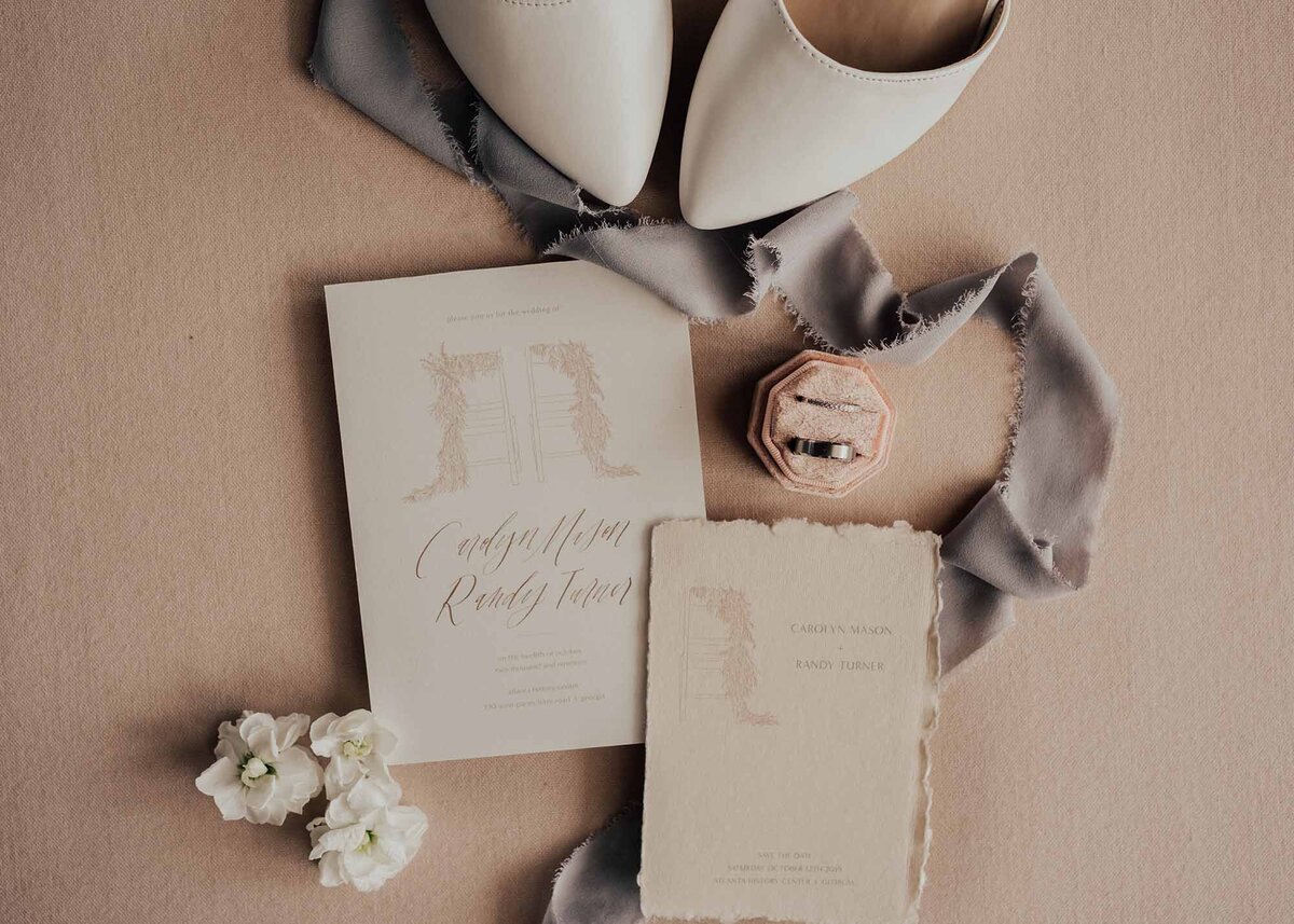 Maddie Rae Photography flat lay of the brides shoes, wedding bands, some flowers, the invitations and there is ribbon going through the image