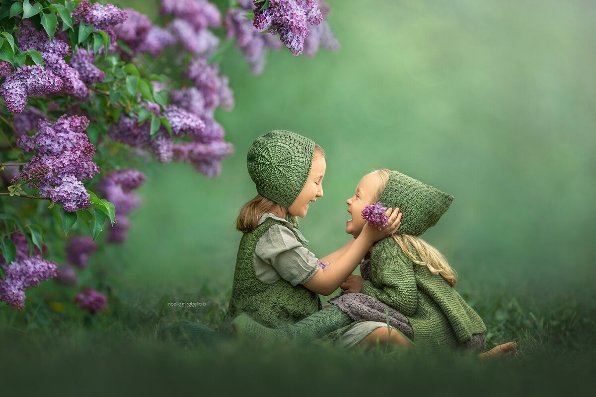 Girls dressed in green knits, laughing and playing under a lilac branch.