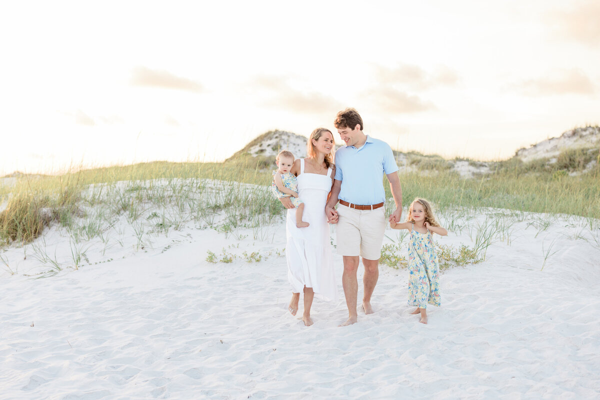 A mom holding a baby with her husband next to her holding the hand of a small child. A sand dune behind them in Watersound Beach Florida.