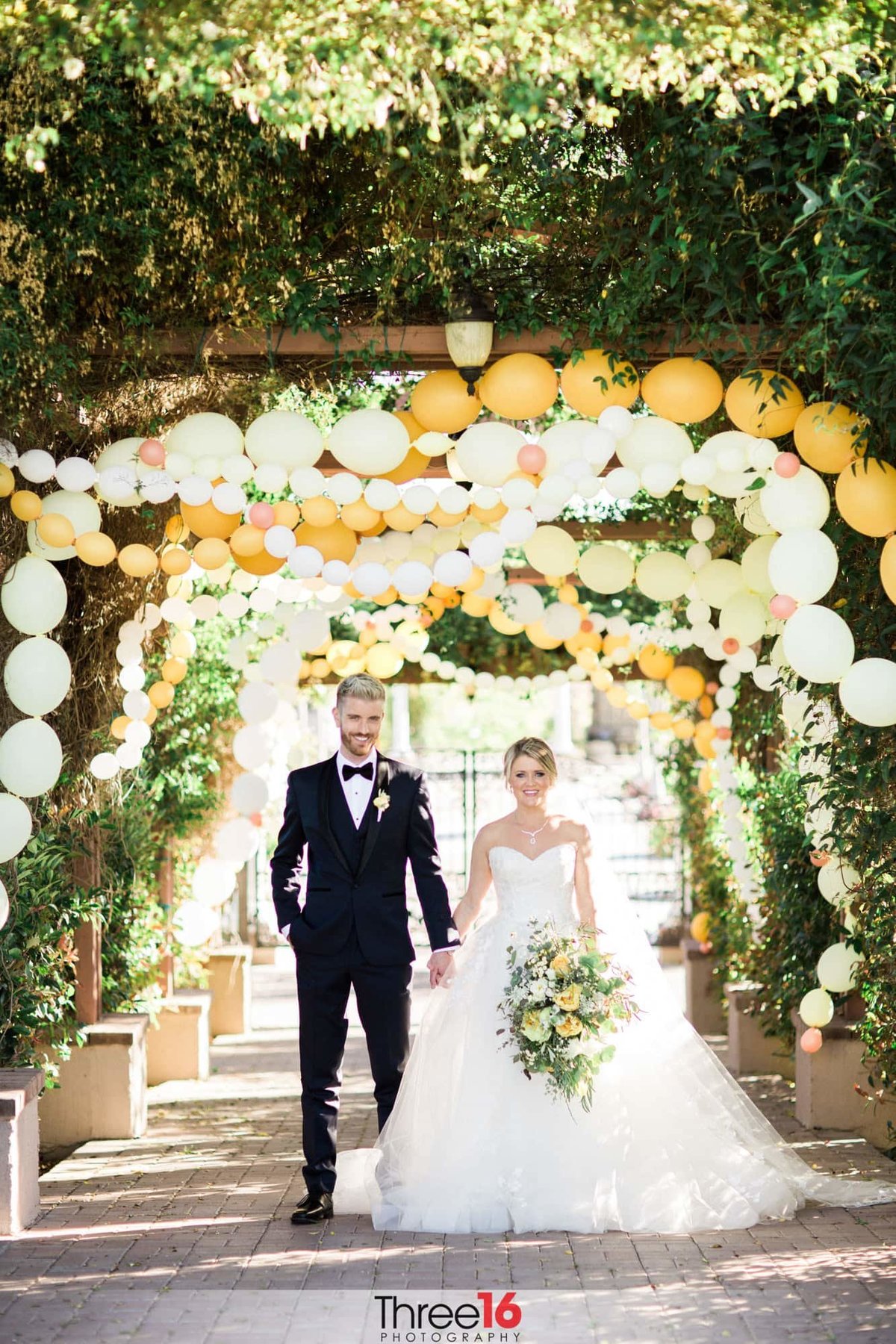 Bride and Groom walk along a path holding hands under a decorative tunnel of balloons