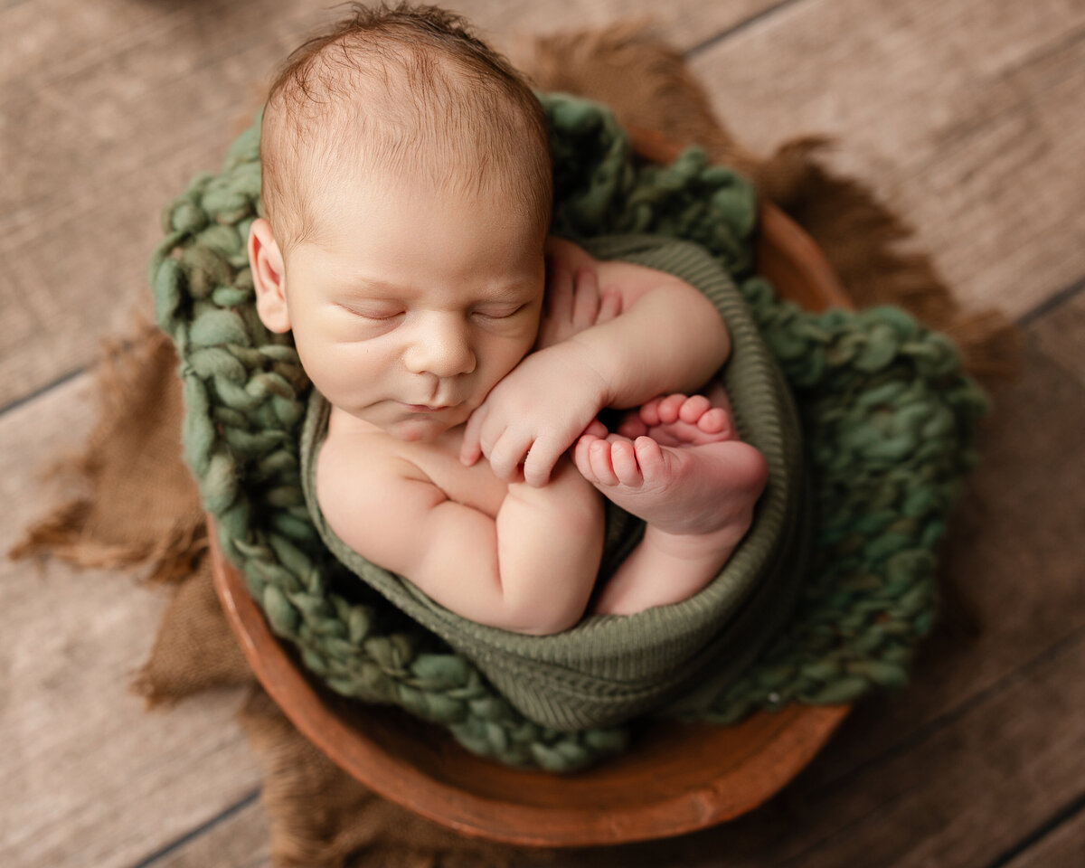 Newborn baby boy curled up in a wooden bowl, wrapped in green fabric with chunky knit layers and warm earth tones. Captured by Inland Empire newborn photographer Kaitlyn Dawn Photography.