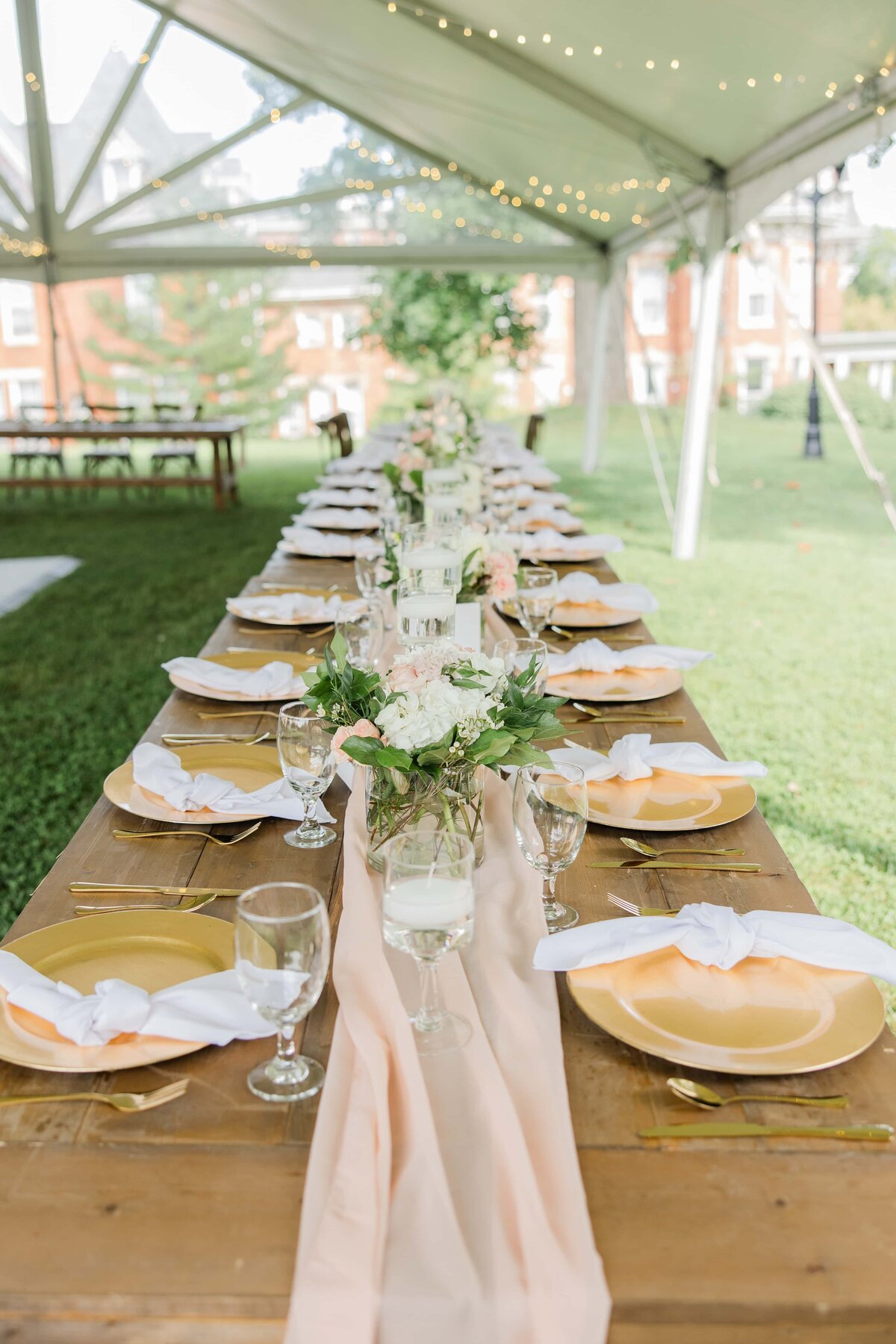Elegantly set dining table for an outdoor Iowa wedding with golden plates, glasses, and floral centerpieces under a tent with string lights.