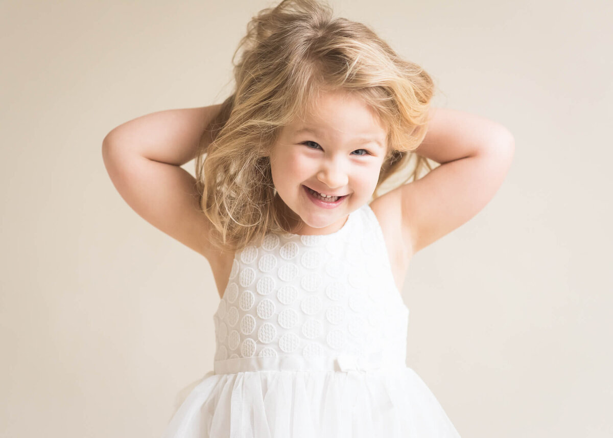 preschool aged girl being silly wearing a white dress with a beige background , light and airy feel