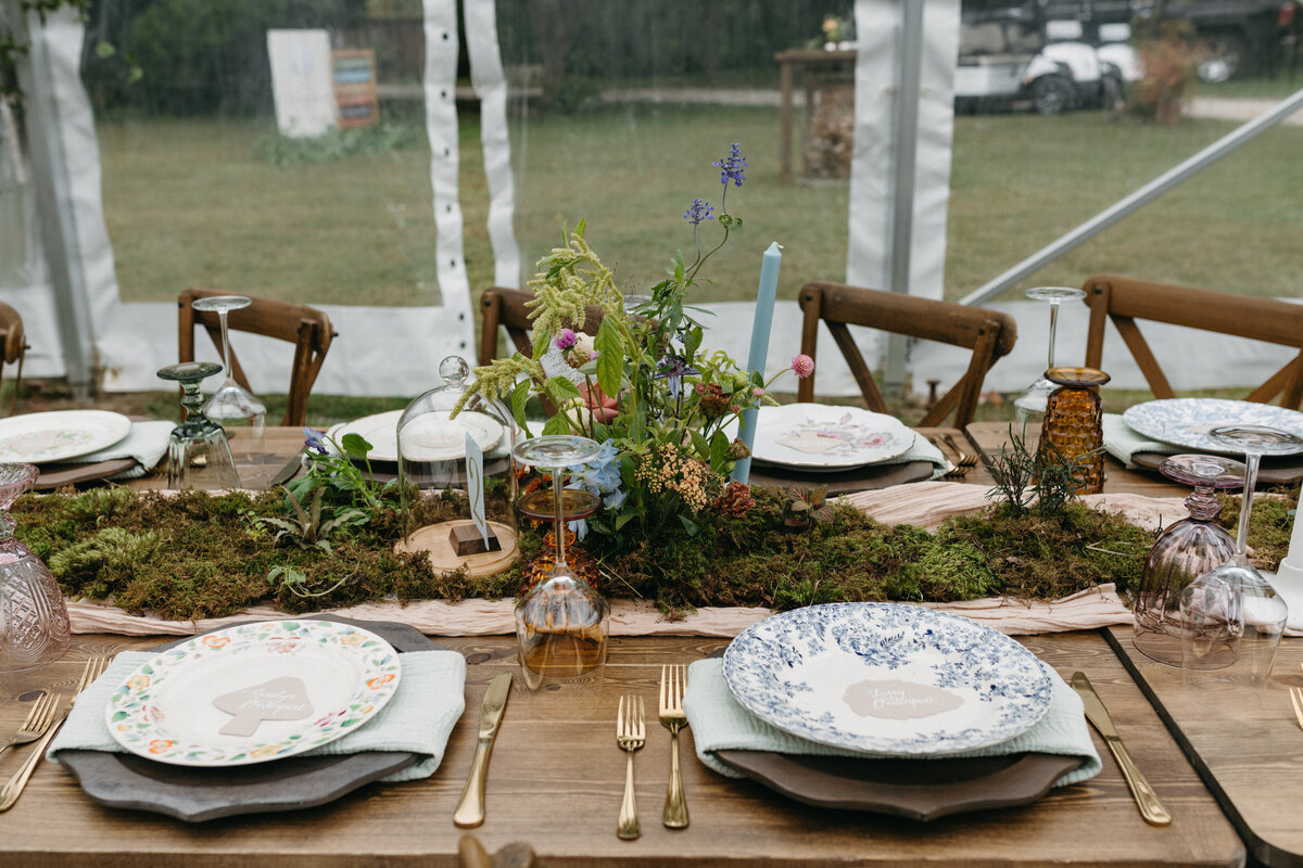 Close-up of a garden-inspired wedding reception table featuring moss table runners, whimsical wildflower centerpieces, pastel taper candles, vintage glassware, and mismatched floral plates arranged by a luxury wedding florist.