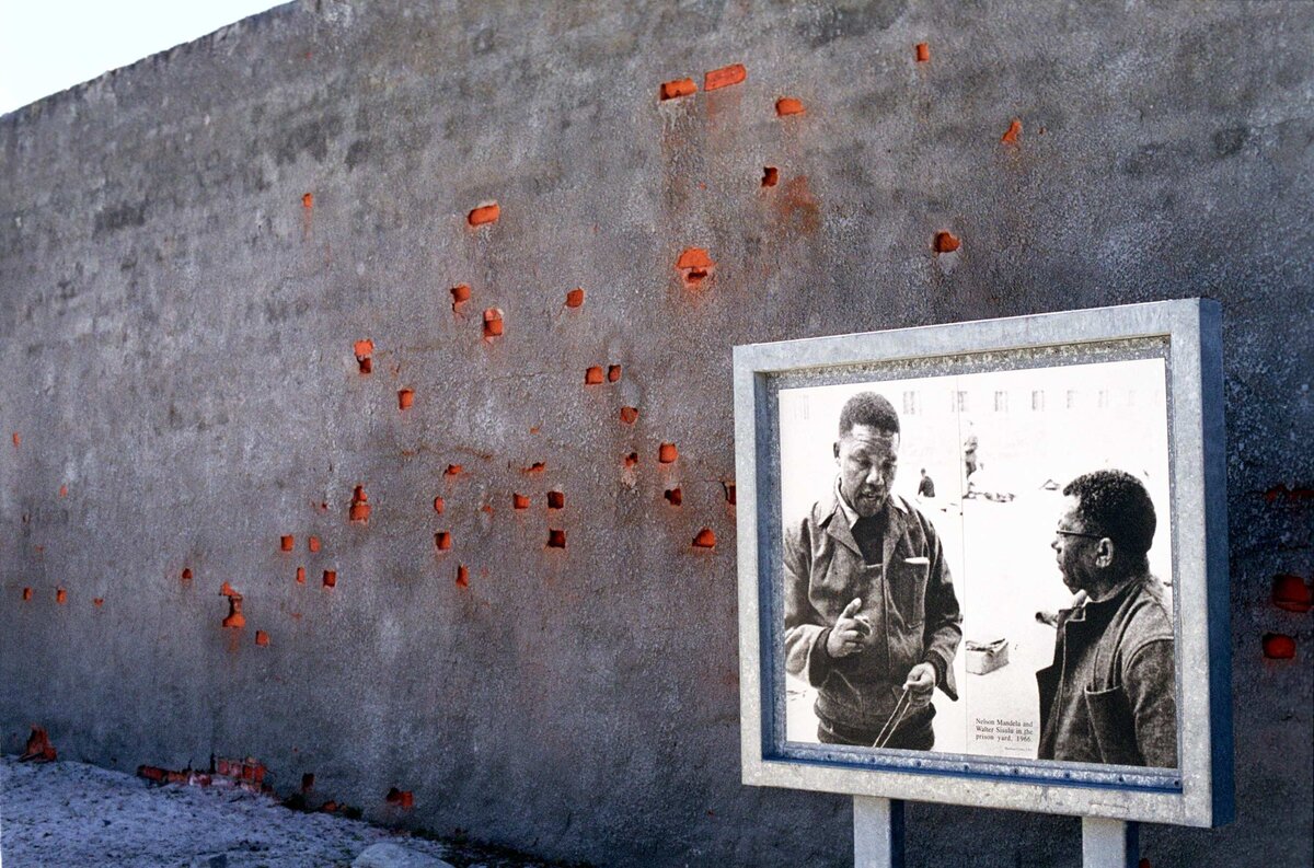 An editorial image of a wall with red bricks bleeding minerals and poster of Nelson Mandela speaking with Steve Biko.