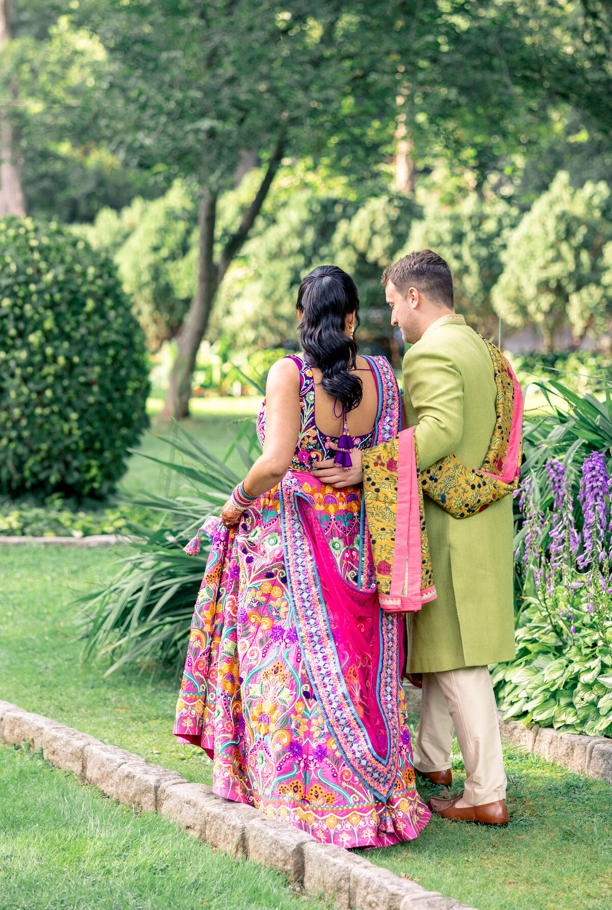 A couple dressed in colorful traditional attire walk arm in arm through a lush garden. The man wears a green jacket, and the woman is in a bright pink sari. They stroll along a stone pathway surrounded by greenery and flowers.