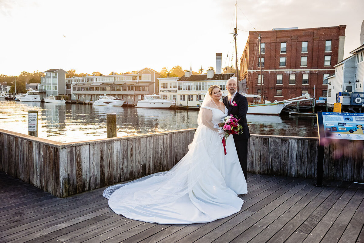 A bride and groom pose on a wooden dock by the water at sunset, with boats and buildings in the background