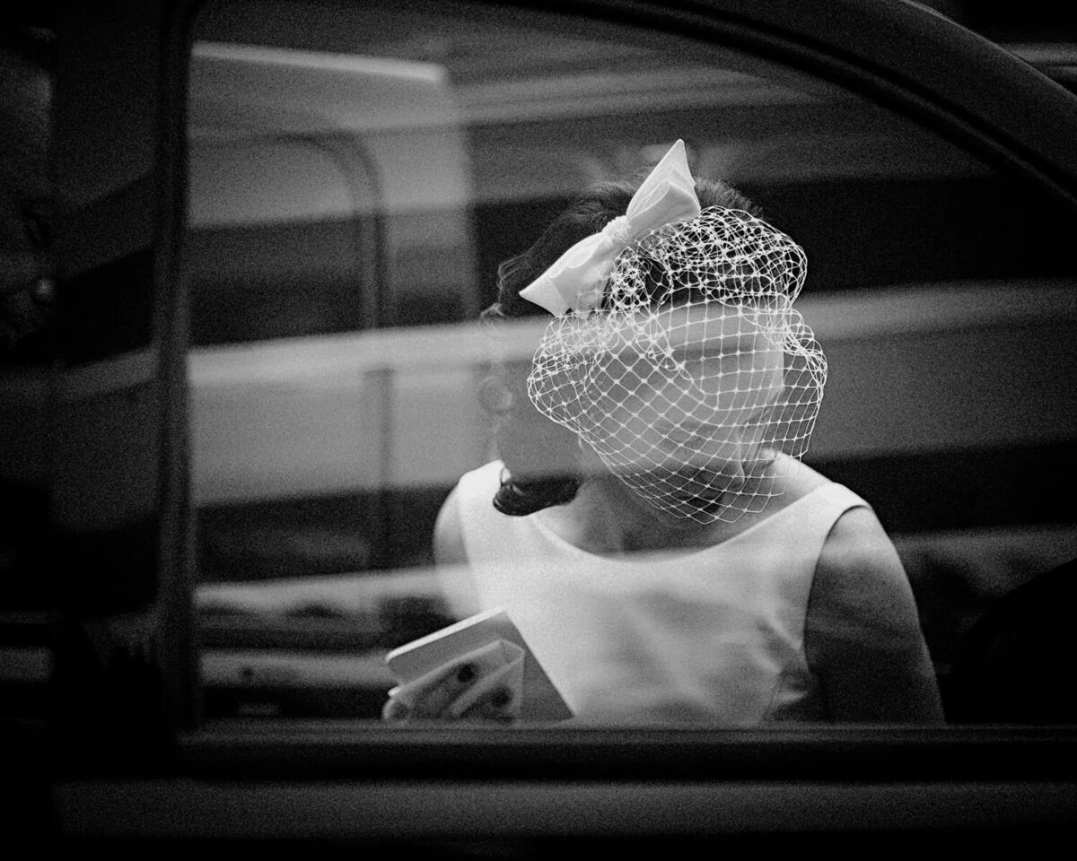 A black and white candid shot of a bride through a car window, wearing a birdcage veil