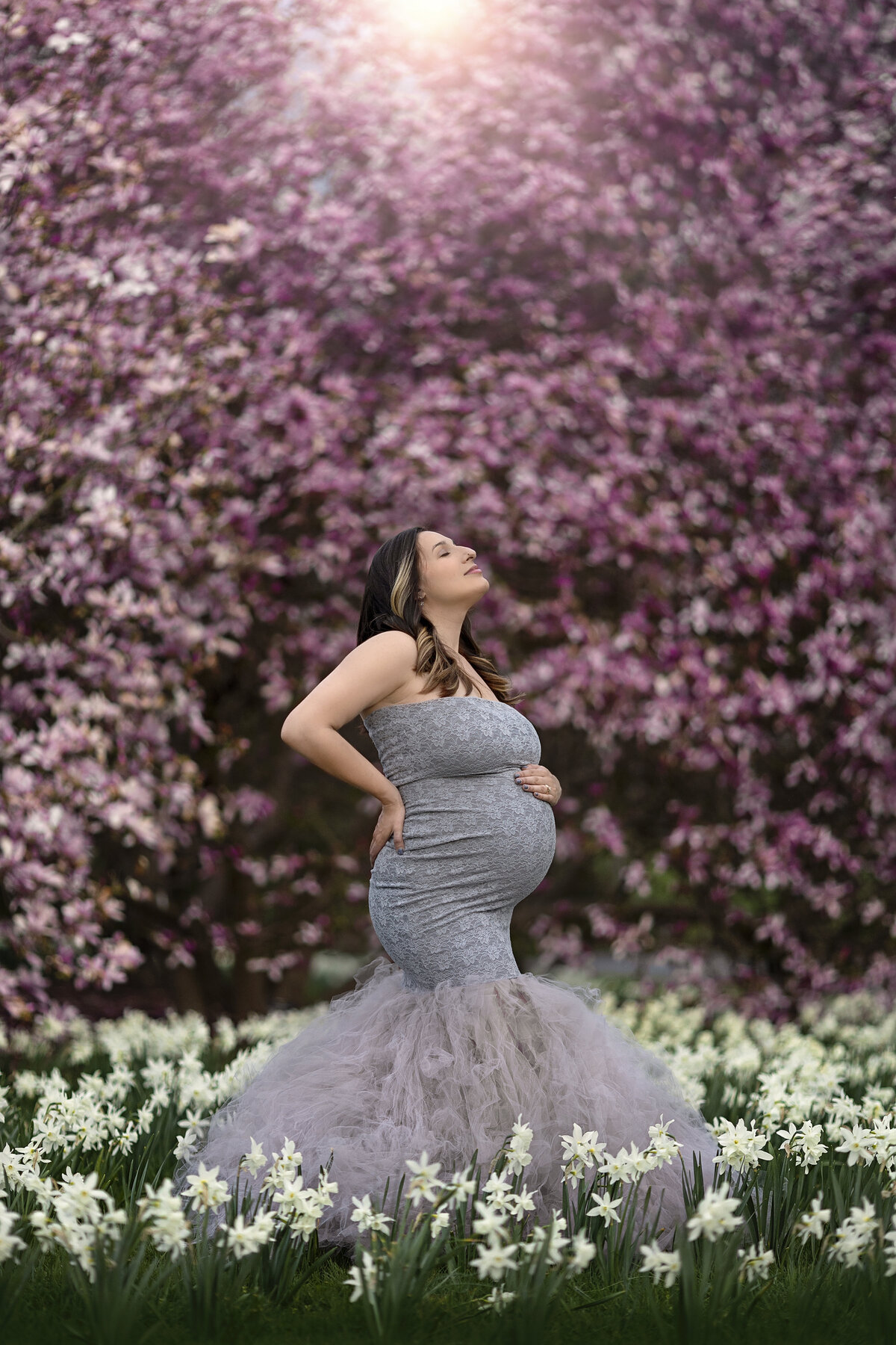 A mother to be stands in a field of wildflowers and blooming trees in a blue/grey maternity gown smiling up to the sun
