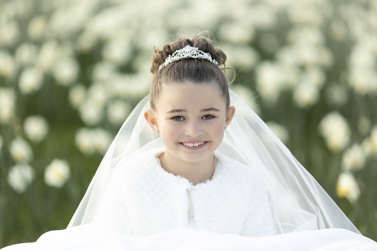 A smiling girl sits in a field of wildflowers in a white dress and tiara for her communion