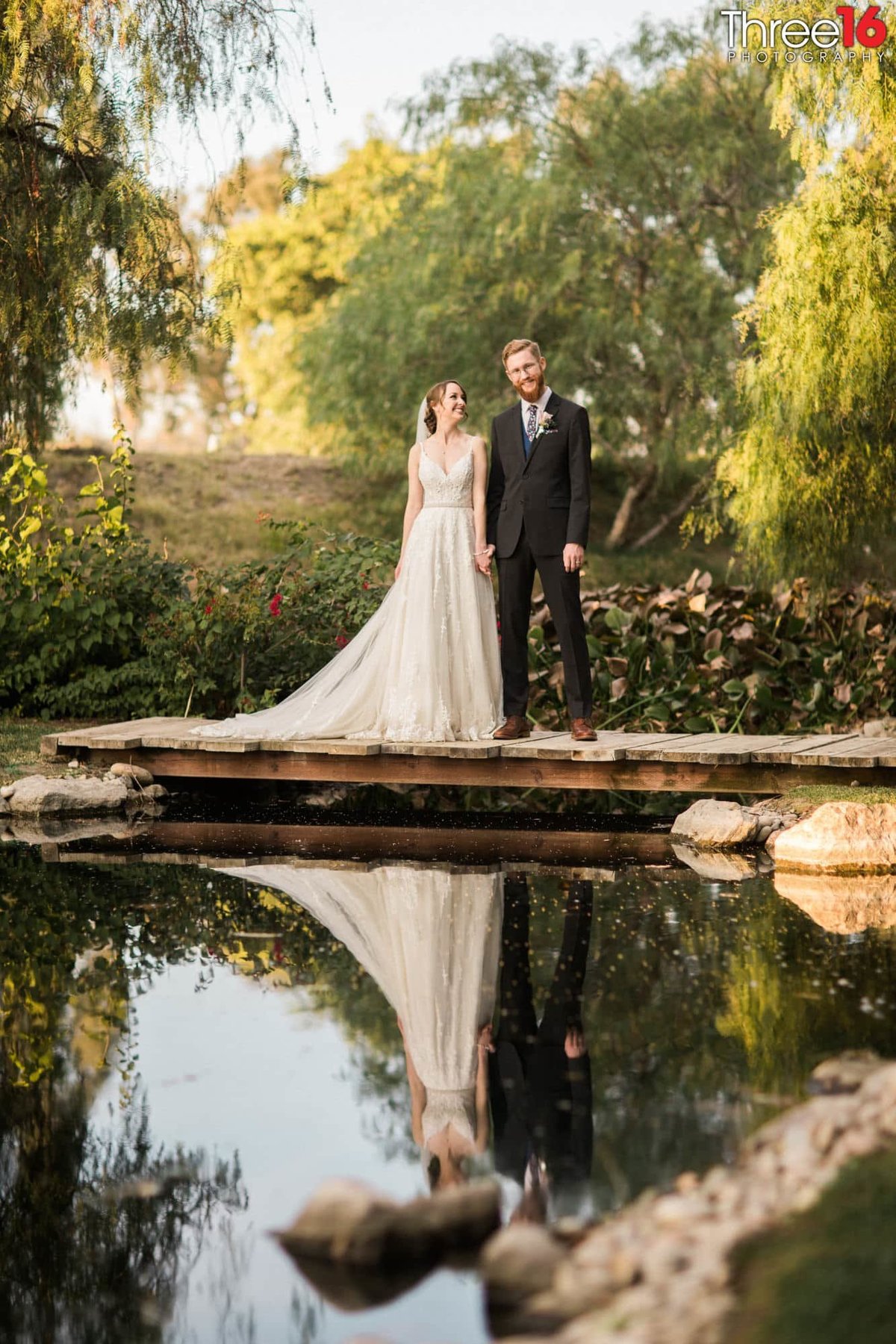 Bride and Groom stand together holding hands on a wooden bridge overlooking a small pond