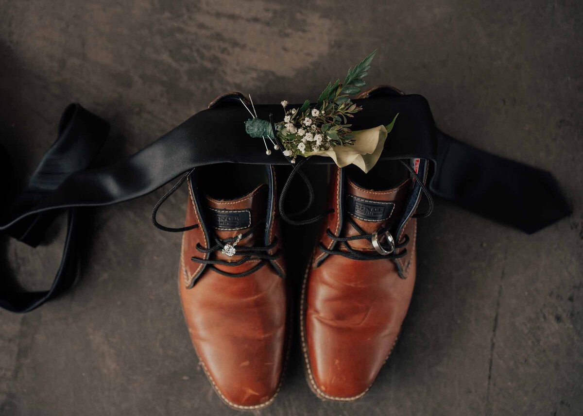 Maddie Rae Photography detail picture of the grooms shoes, his tie is laying untied over his shoes, the boutonniere is resting on the tie, and the bride and grooms rings are looped in the laces of each shoe