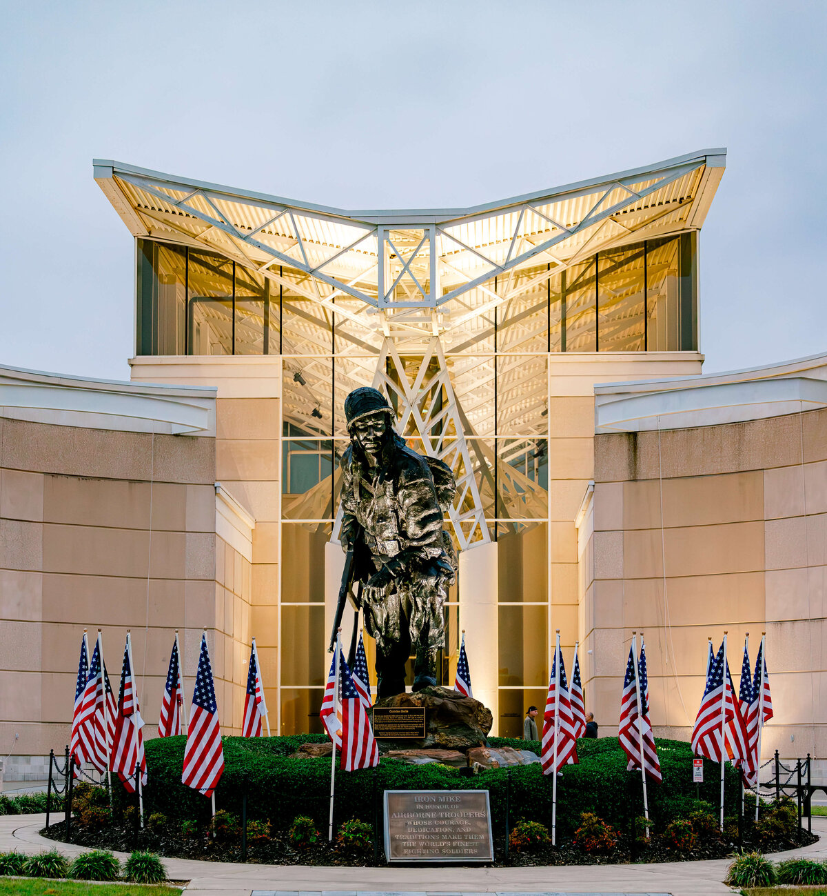 An event photographer in Fayetteville, NC captures the striking image of a large soldier statue standing in front of a modern building with a unique roof design. American flags line the scene, and a plaque is visible at the base. The overcast sky adds to the solemn atmosphere.