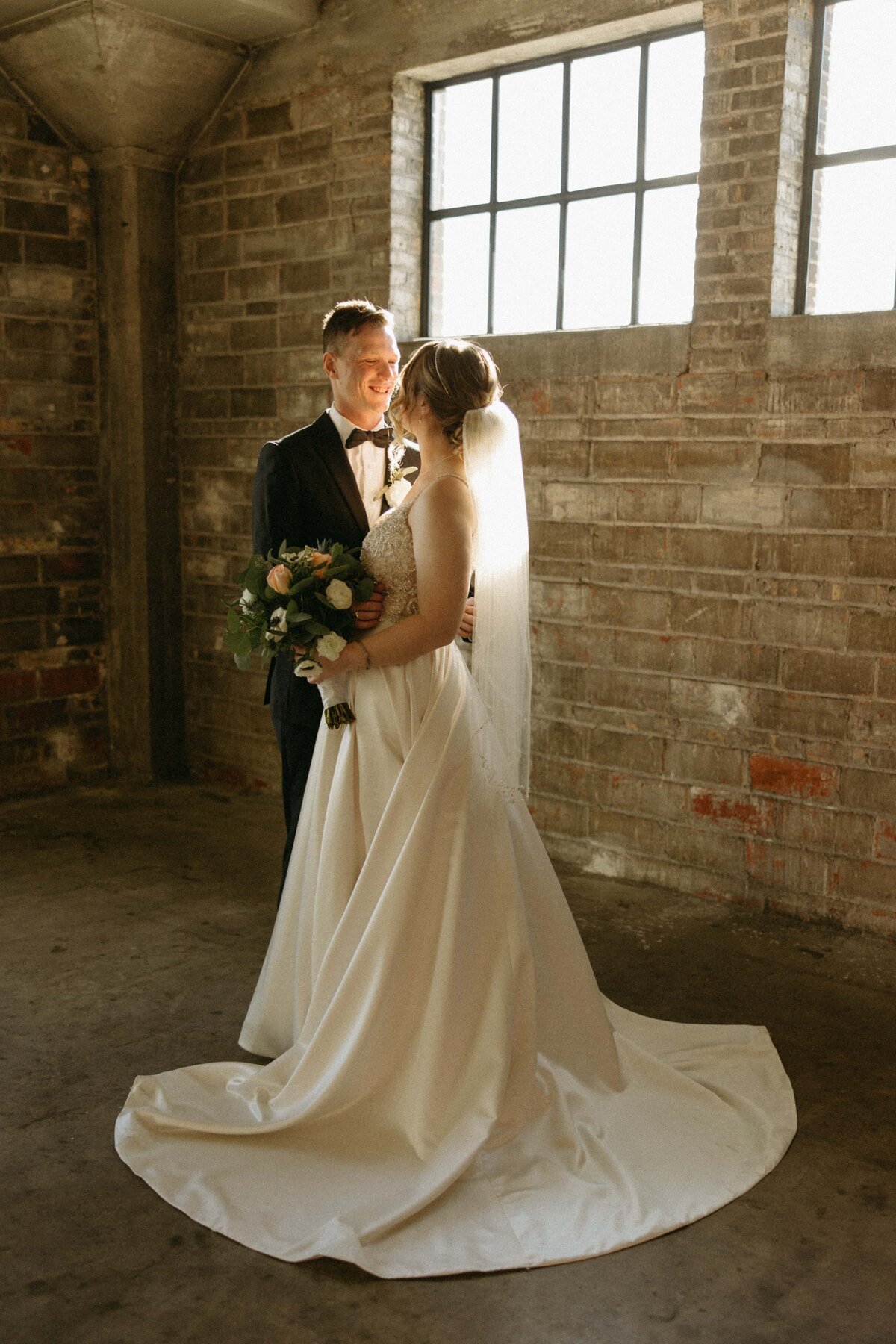 A bride and groom smiling at each other in a sunlit, rustic brick room, the bride holding a bouquet while wearing an elegant white dress with a long train, perfectly arranged by their wedding coordinator in