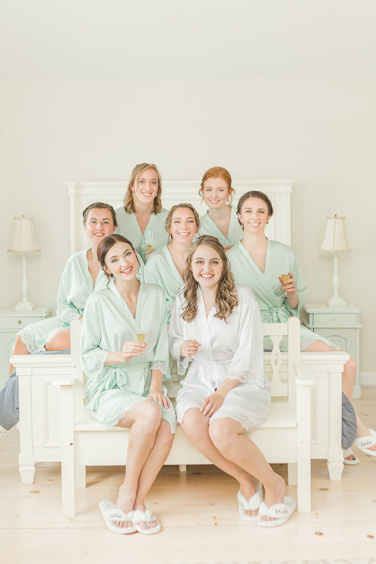 A bride and her bridesmaids gather for a formal getting ready portrait. The bride is in a white robe and her bridesmaids in pale green robes. They are all holding glasses of champagne, sitting on a bed and smiling at the camera.