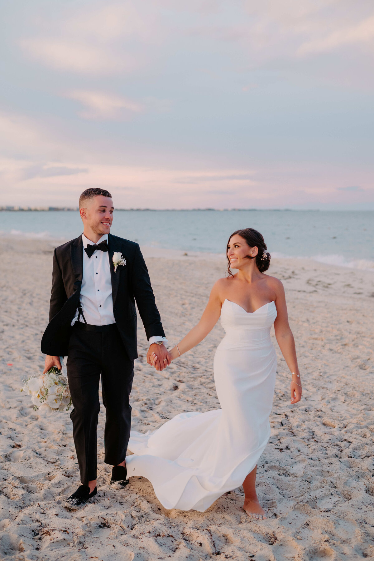 A couple in wedding attire walks barefoot on a beach at sunset. The groom wears a black tuxedo, and the bride wears a flowing white gown. They hold hands, looking joyful.