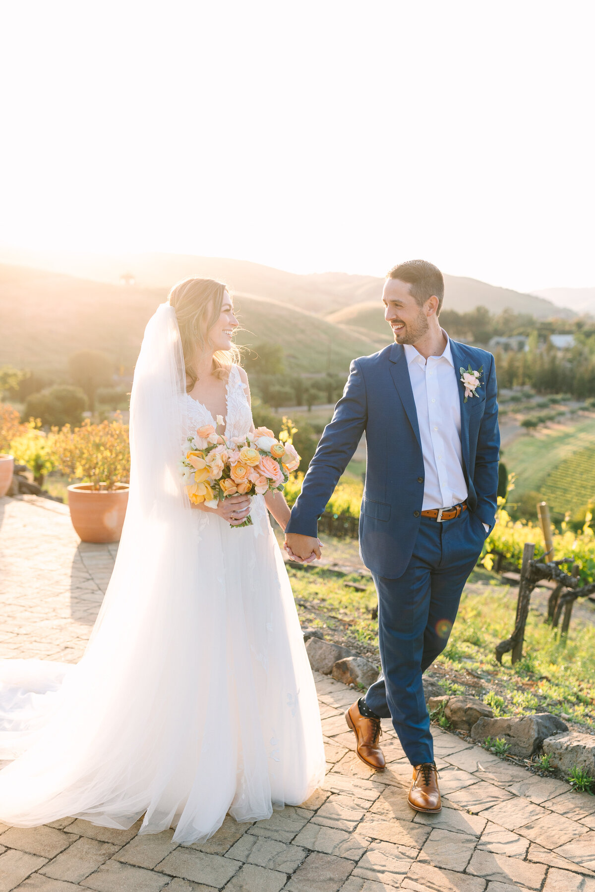 bride with floral bouquet holding hands with groom in napa valley vineyard at golden hour.