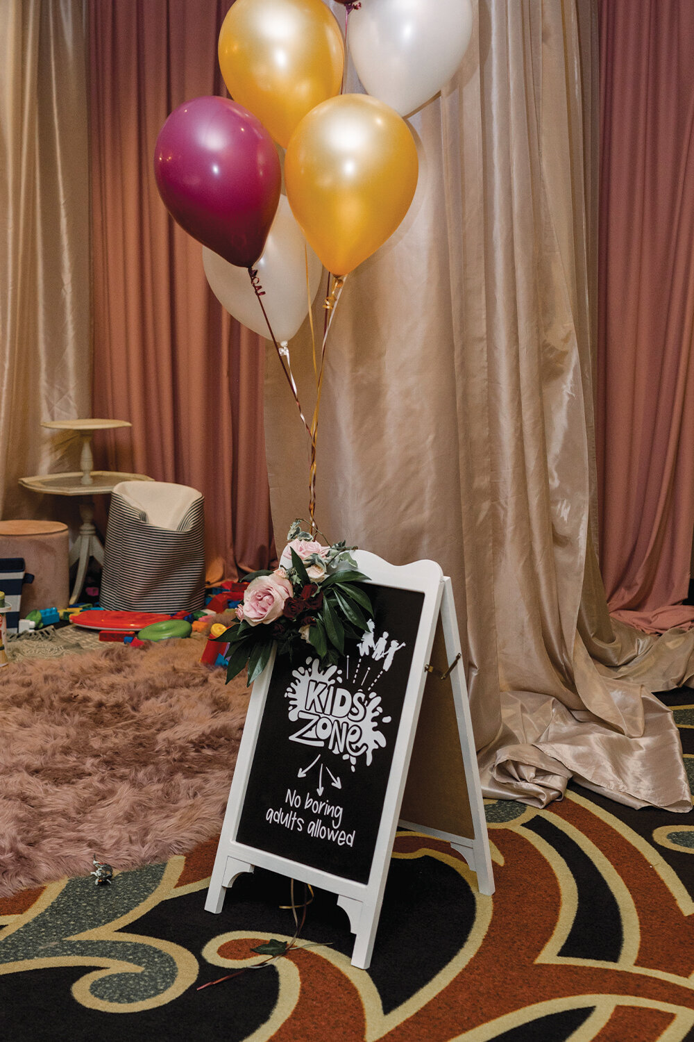 Chalkboard sign reading ‘Kids Zone. No Boring Adults Allowed’ at a 50th wedding anniversary celebration, adorned with burgundy and gold balloons and cascading floral elements. This playful and inviting setup adds a touch of whimsy to the event at The Northern Hotel in Billings, MT