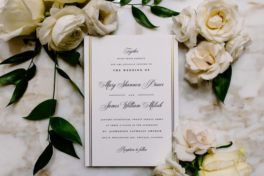 Elegant Chicago wedding invitation surrounded by white roses and green leaves lay on marble countertop.