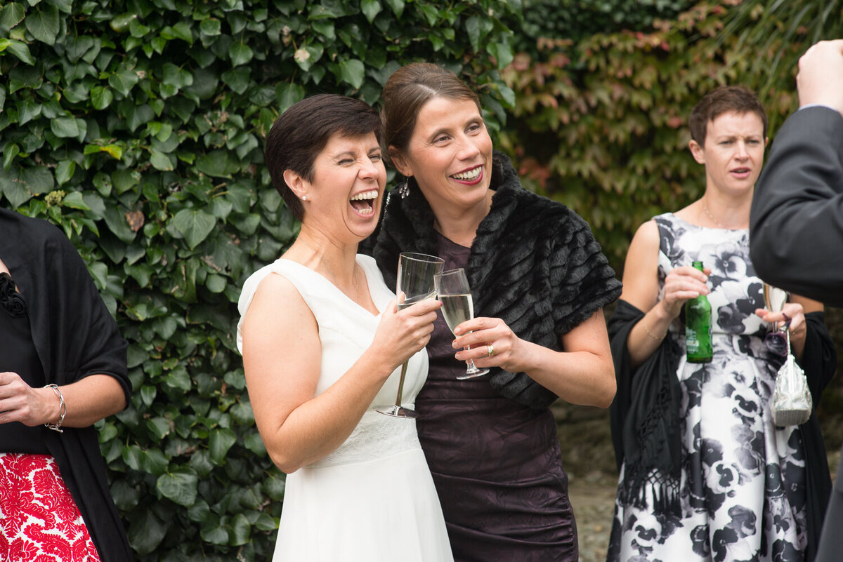 Bride in white laughing with guest in black, holding champagne