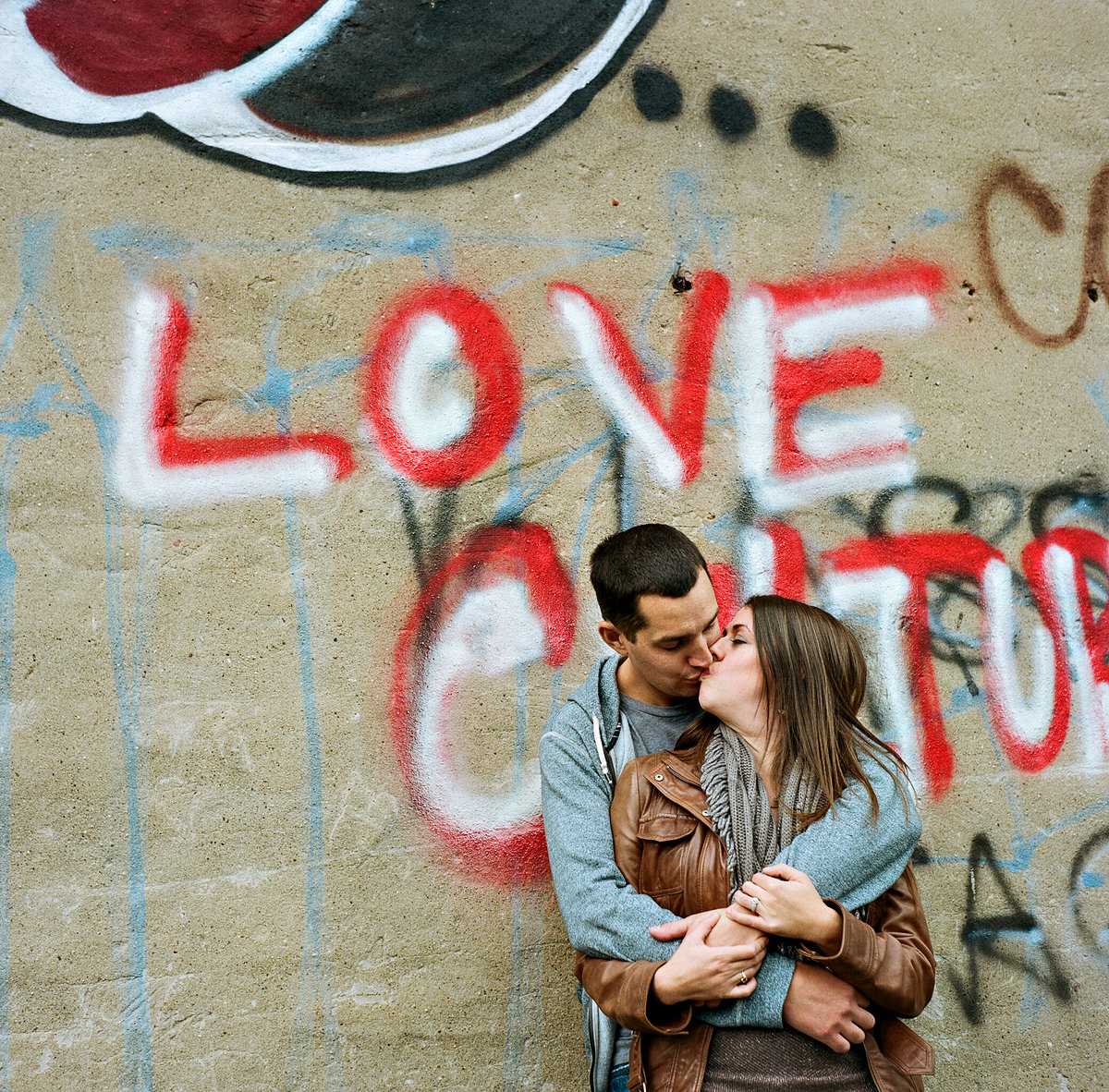 A couple kiss with the word love written on the wall behind them.