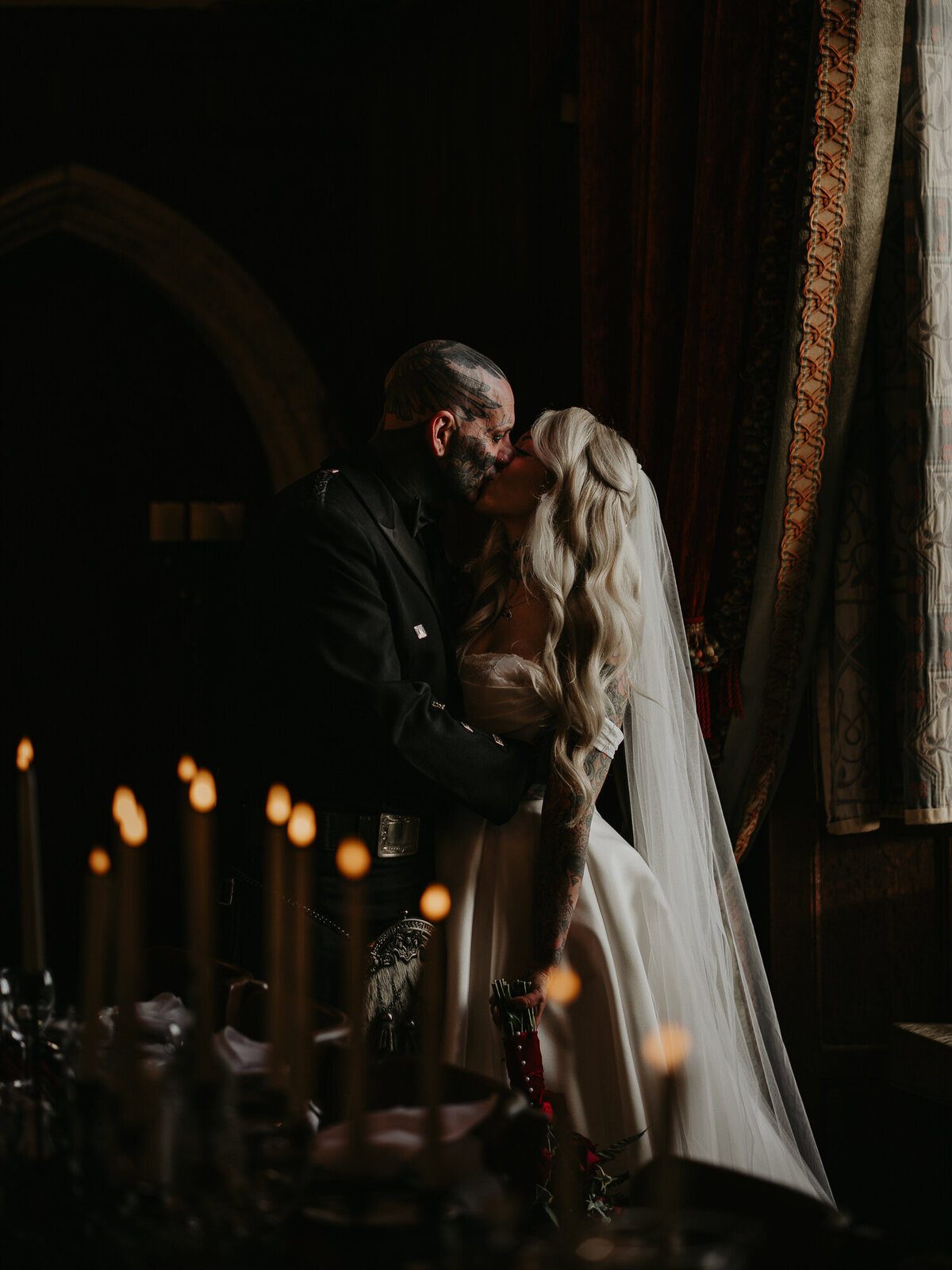 A tattooed bride and groom kiss in the window light at at Lympne Castle in Kent.