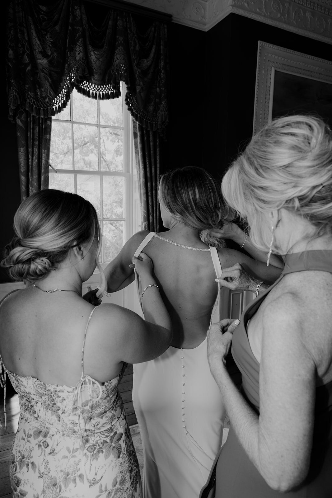 Bridesmaid and mother of the bride helping bride into wedding dress. Photo shows all three from behind standing in William Aiken House Bridal suite.