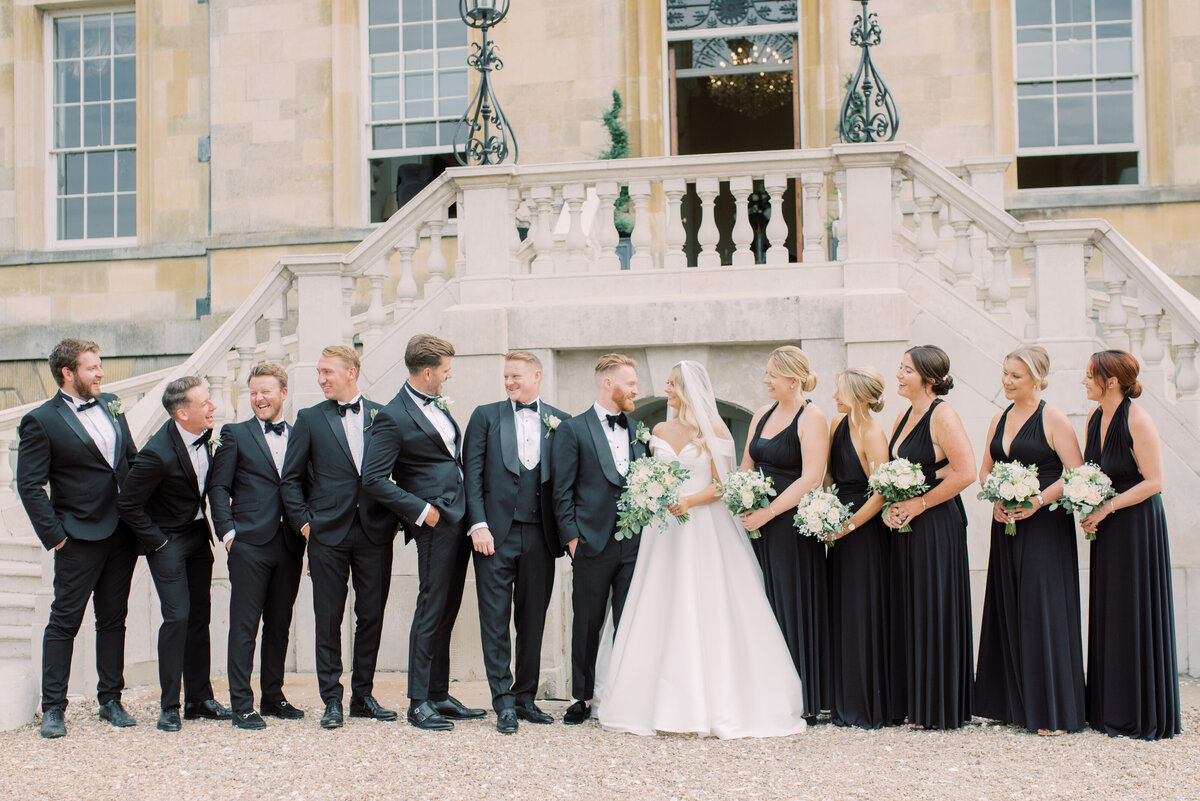 Bridal party lined up outside the steps at botleys mansion surrey, the bridesmaids are wearing black dresses and holding white bouqetsm the groomsmen are in black tuxedos