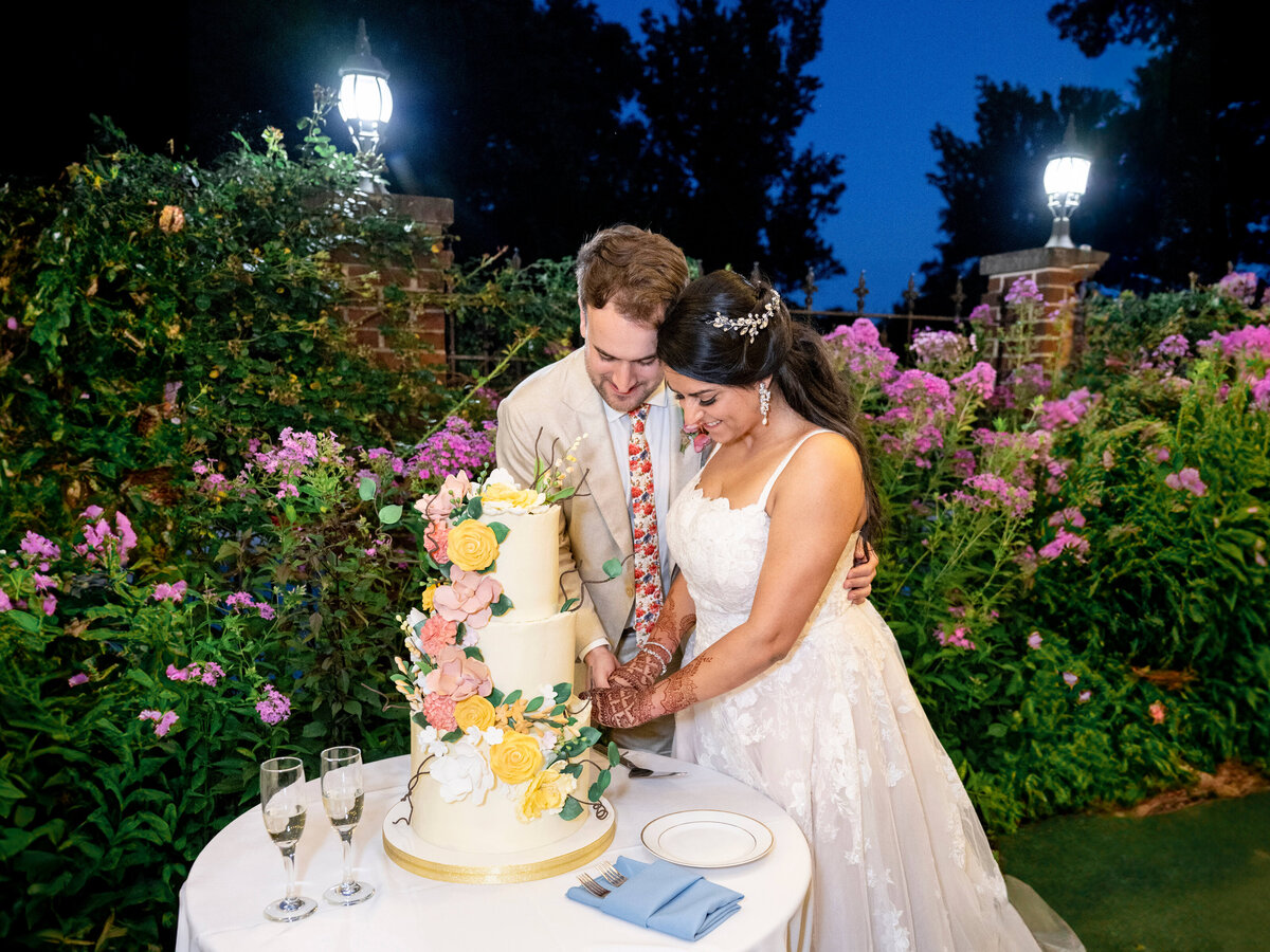 A bride and groom are cutting a white and yellow tiered wedding cake adorned with flowers. They are smiling and surrounded by lush greenery and purple flowers, with two glasses of champagne on the table. Its nighttime with lit lamps in the background.