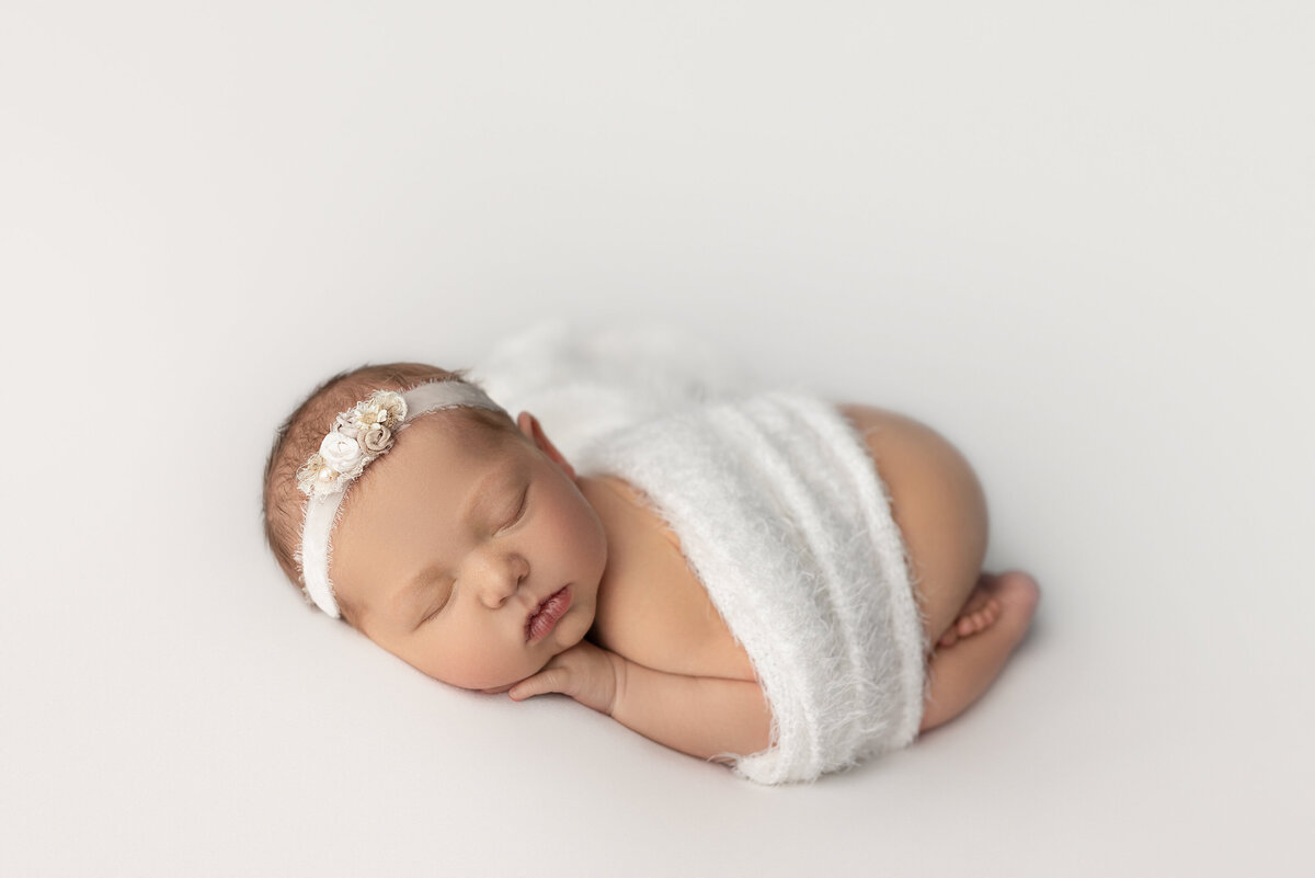 Fine art newborn photos captured by best New Jersey newborn photographer Katie Marshall. Side profile image. Baby girl is sleeping on her belly with her legs curled underneath her. Her head is turned toward the camera. Her head is resting atop of her hand. Baby is sleeping on white stretch fabric.