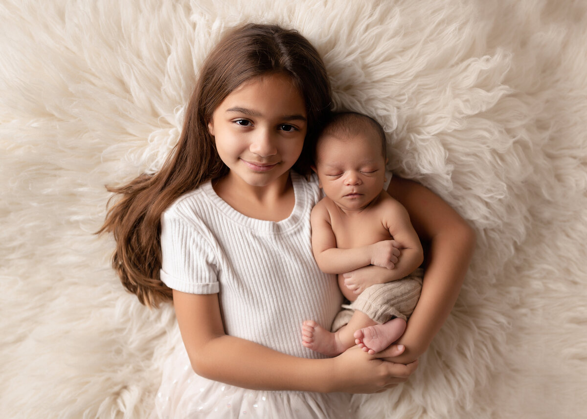 Sibling newborn photo in West Palm Beach and Boca Raton photo studio - aerial image. Big sister is holding baby brother atop of a long faux fur rug. Big sister has her long brown hair draped over her shoulder. Baby brother is wearing cream colored knit pants and is sleeping with his head resting on big sister's shoulder.