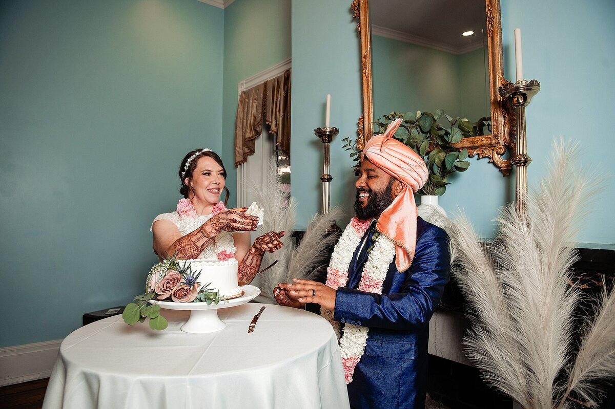The bride, wearing a rhinestone headband and mendhi on her hands feeds the groom cake at their wedding reception. The groom is wearing a cobalt suit, a white and pink varmala and a pink turban. The small white wedding cake is on a white footed stand at Ravenswood Mansion.
