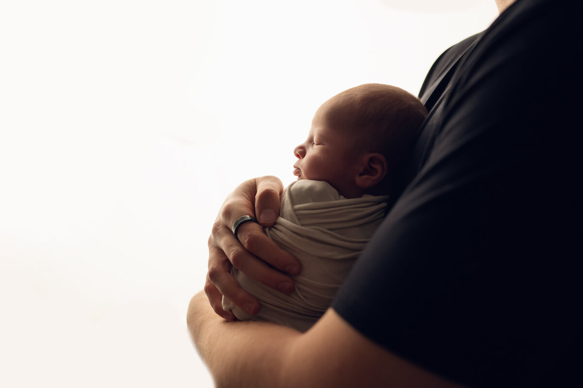 Profile portrait of a newborn baby wrapped in his father's arms. Only shoulder and arm of dad is shown holding swaddled infant to his chest.