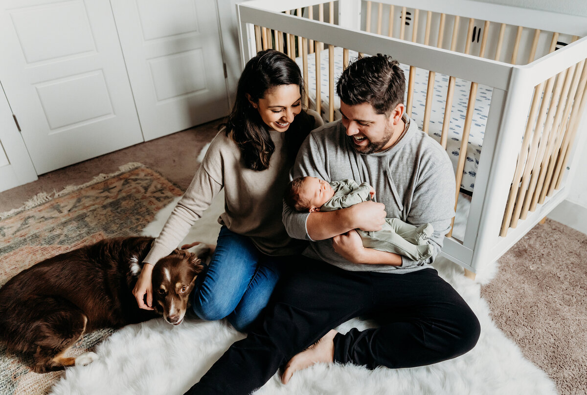 Newborn Photographer, mom and dad sit in the nursery excitedly smiling at their baby, their dog sits beside them
