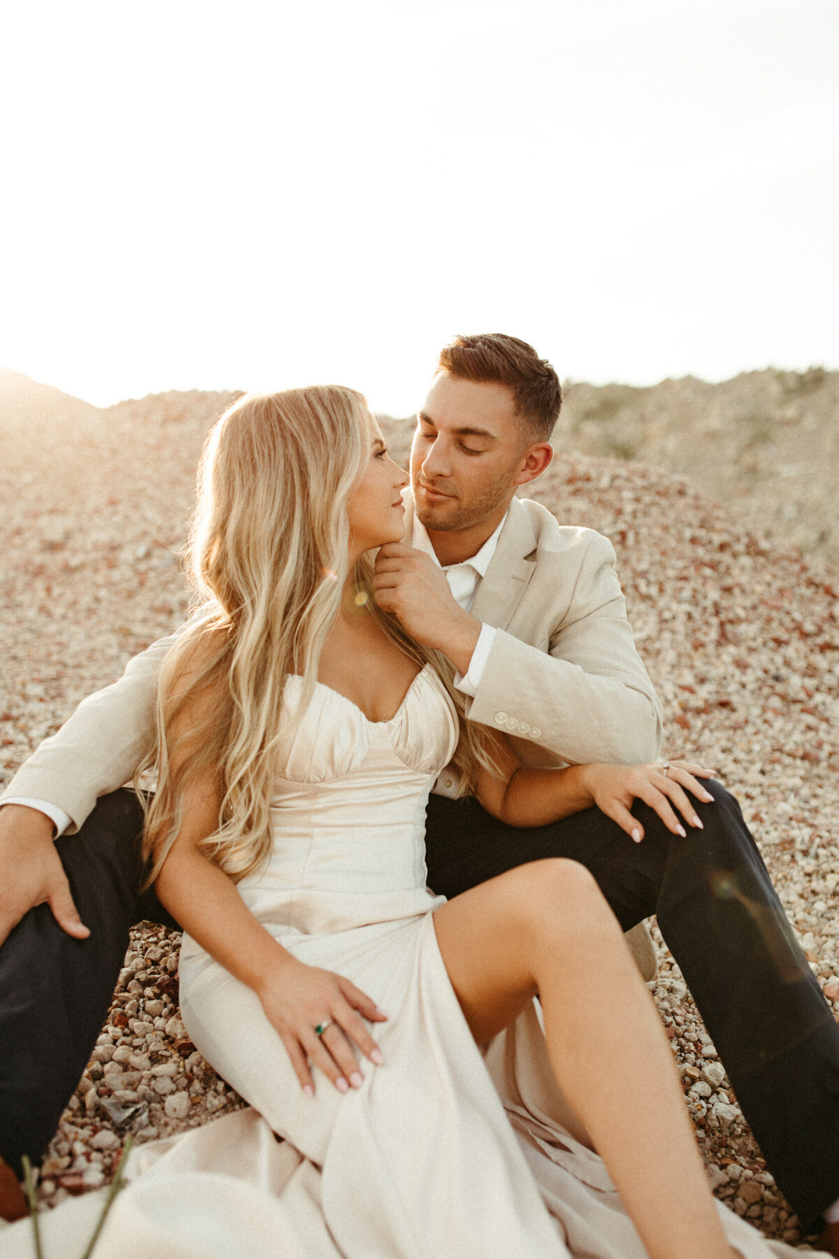 A girl in a silk champagne colored dress is sitting down between her fiancé's legs while he pulls her in for a kiss.