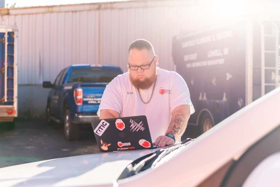 A man with a beard sits outdoors using a laptop with stickers, near vehicles in a sunny setting.