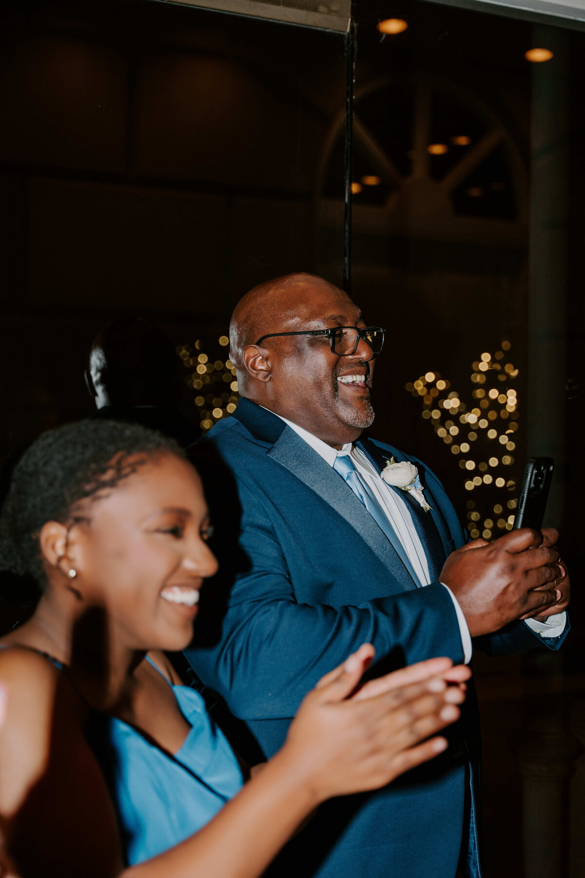 A joyous scene at an event: A man in a blue suit smiles broadly and holds a phone, while a woman in a blue dress claps. Warm lights twinkle in the background.